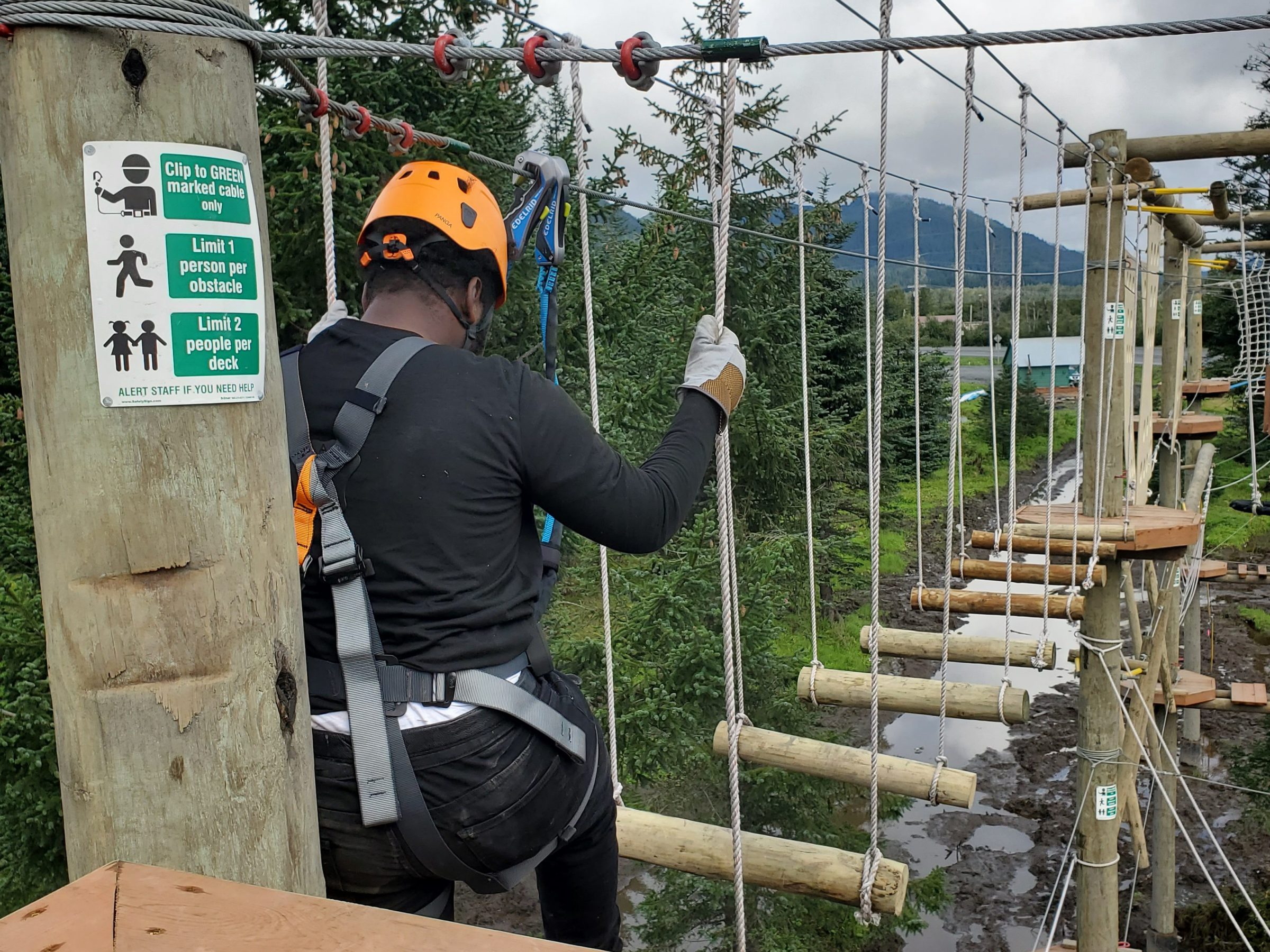 Person in safety gear crossing a treetop adventure course with ropes and logs.