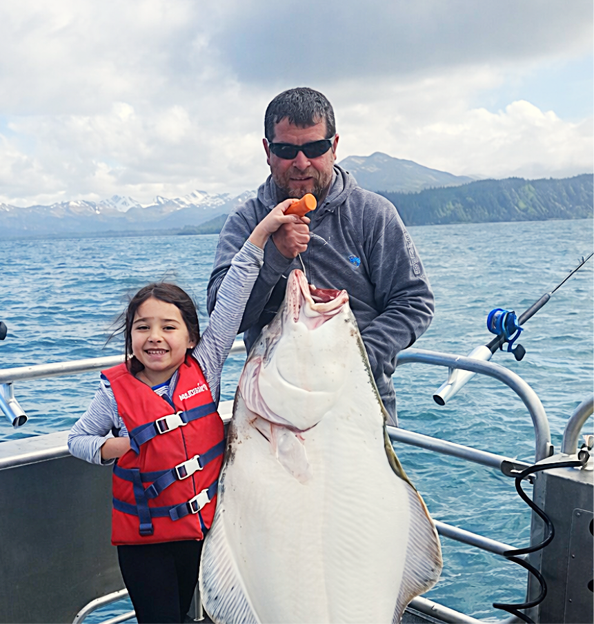 Man and girl on boat holding large fish with mountains in background.