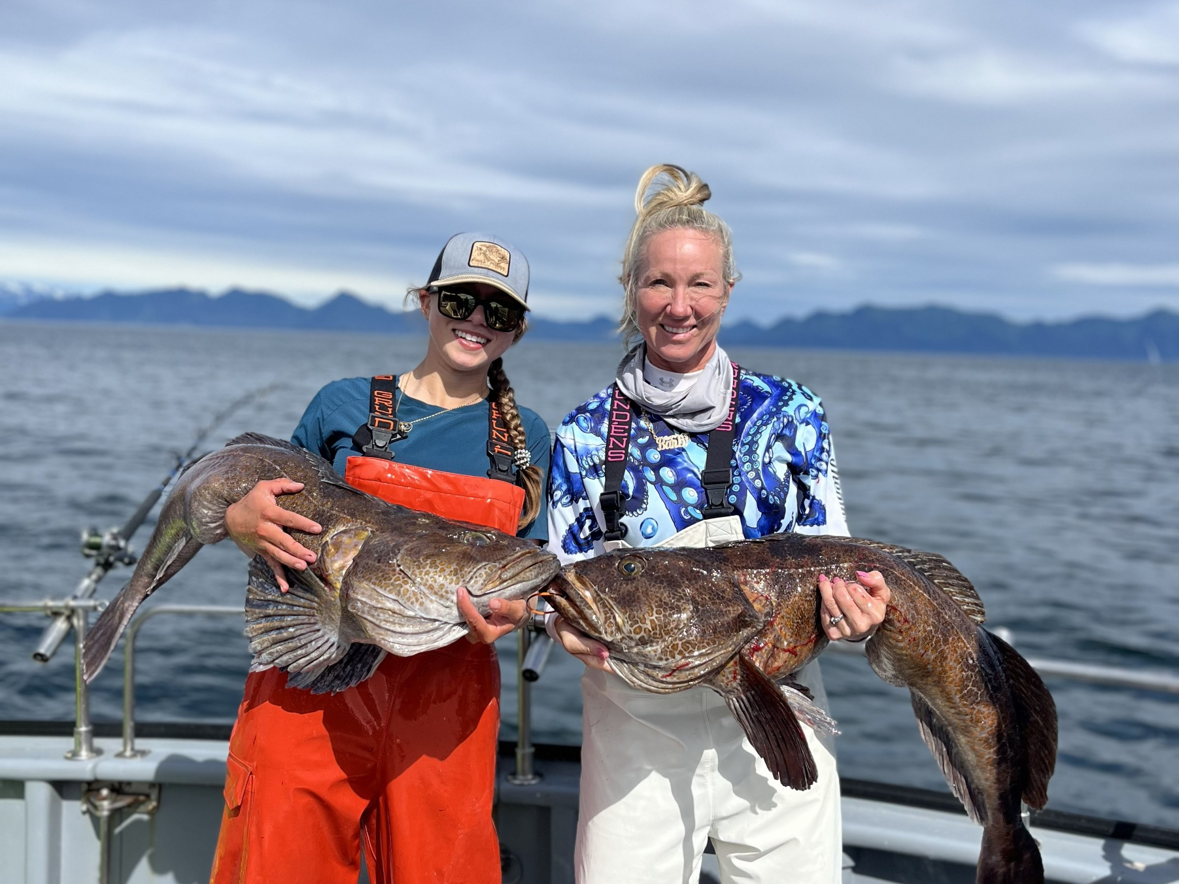 Two people on a boat holding large fish with a mountainous background.