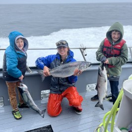 Three people on a boat holding caught fish, smiling with ocean in background.