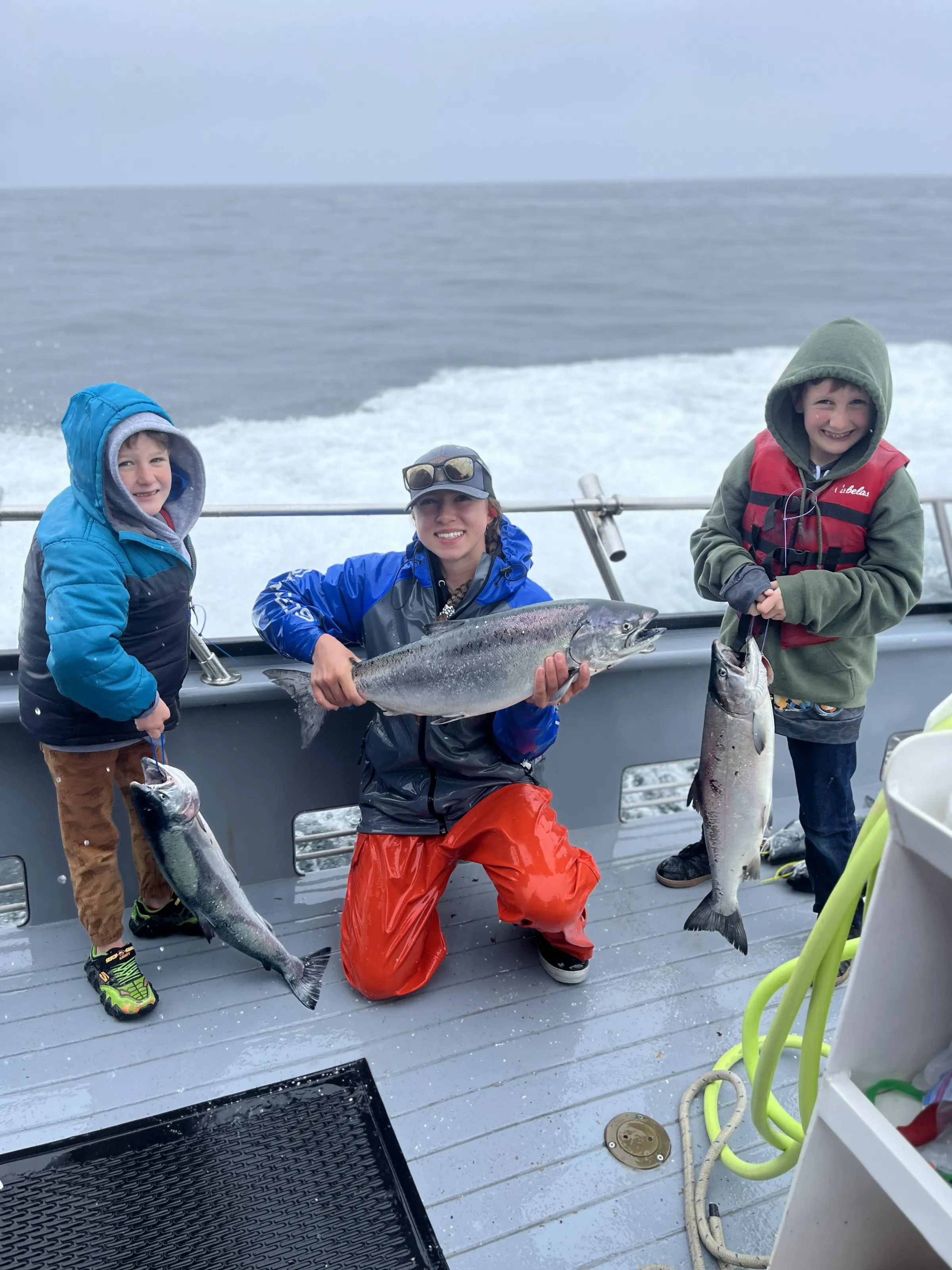 Three people on a boat holding caught fish, smiling with ocean in background.