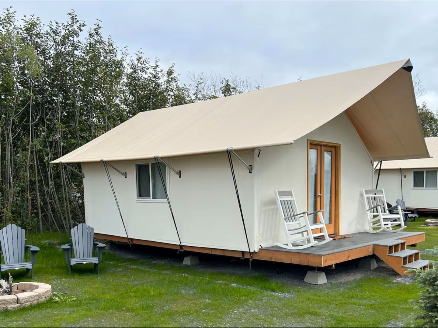 Canvas tent cabin with rocking chairs on porch next to a fire pit and chairs in a grassy area.
