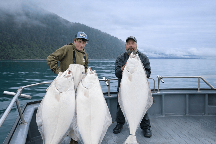 Two people on a boat holding large white fish against a misty forested background.
