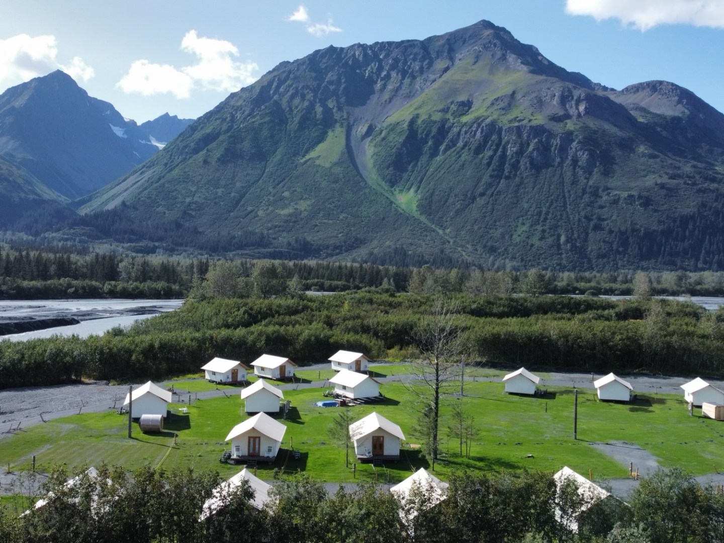 Tents on a grassy field with a mountain backdrop and river in the foreground.
