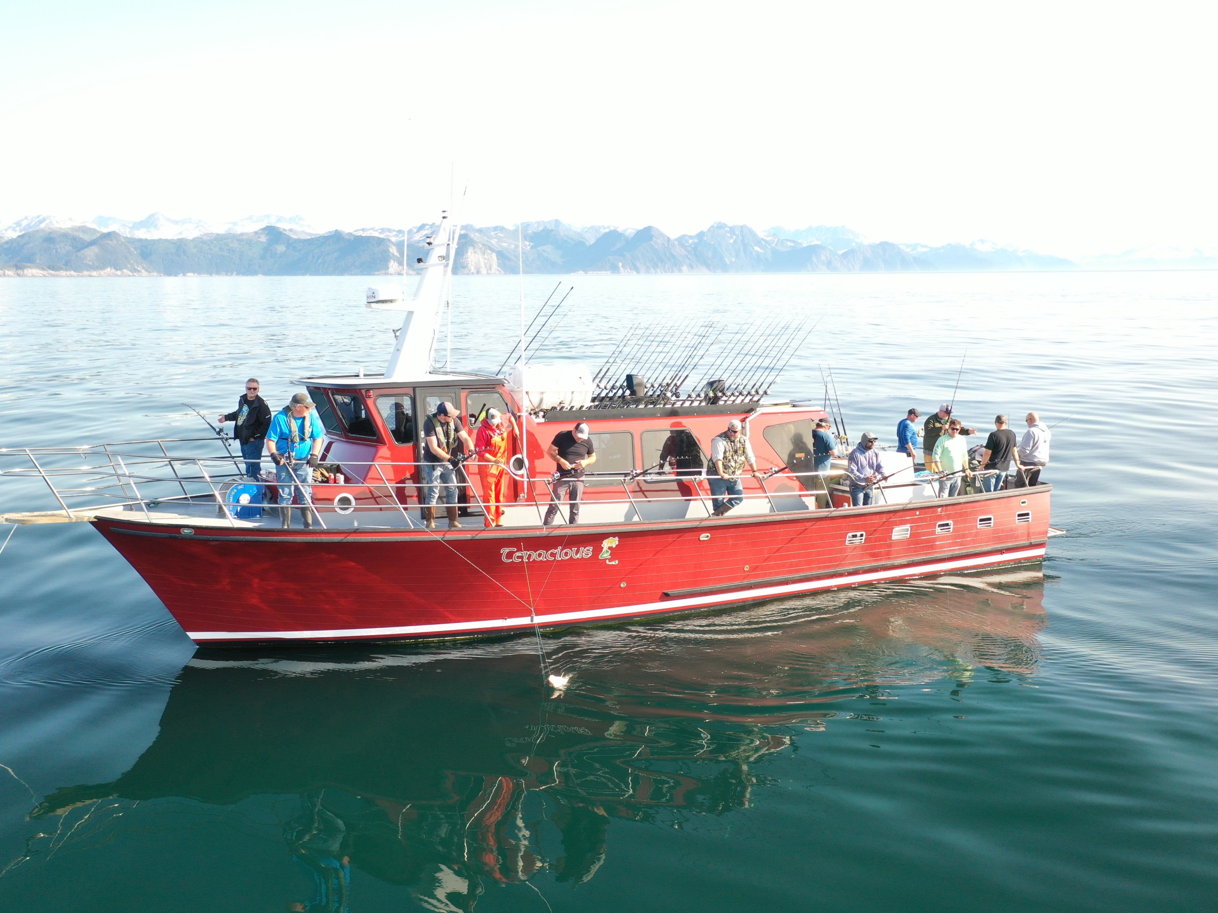 Red boat with people fishing in calm waters, mountains in background, clear sky.