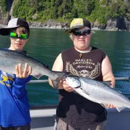 Two people on a boat holding large fish, wearing caps and sunglasses, with a forested shore in the background.