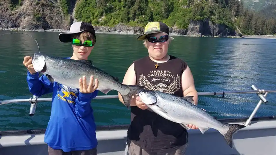 Two people on a boat holding large fish, wearing caps and sunglasses, with a forested shore in the background.