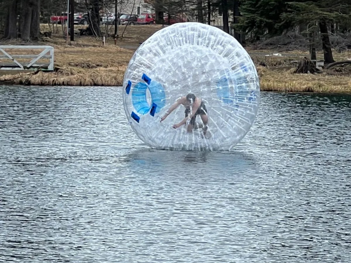 Person inside a large inflatable ball floating on a lake near a forested shore.