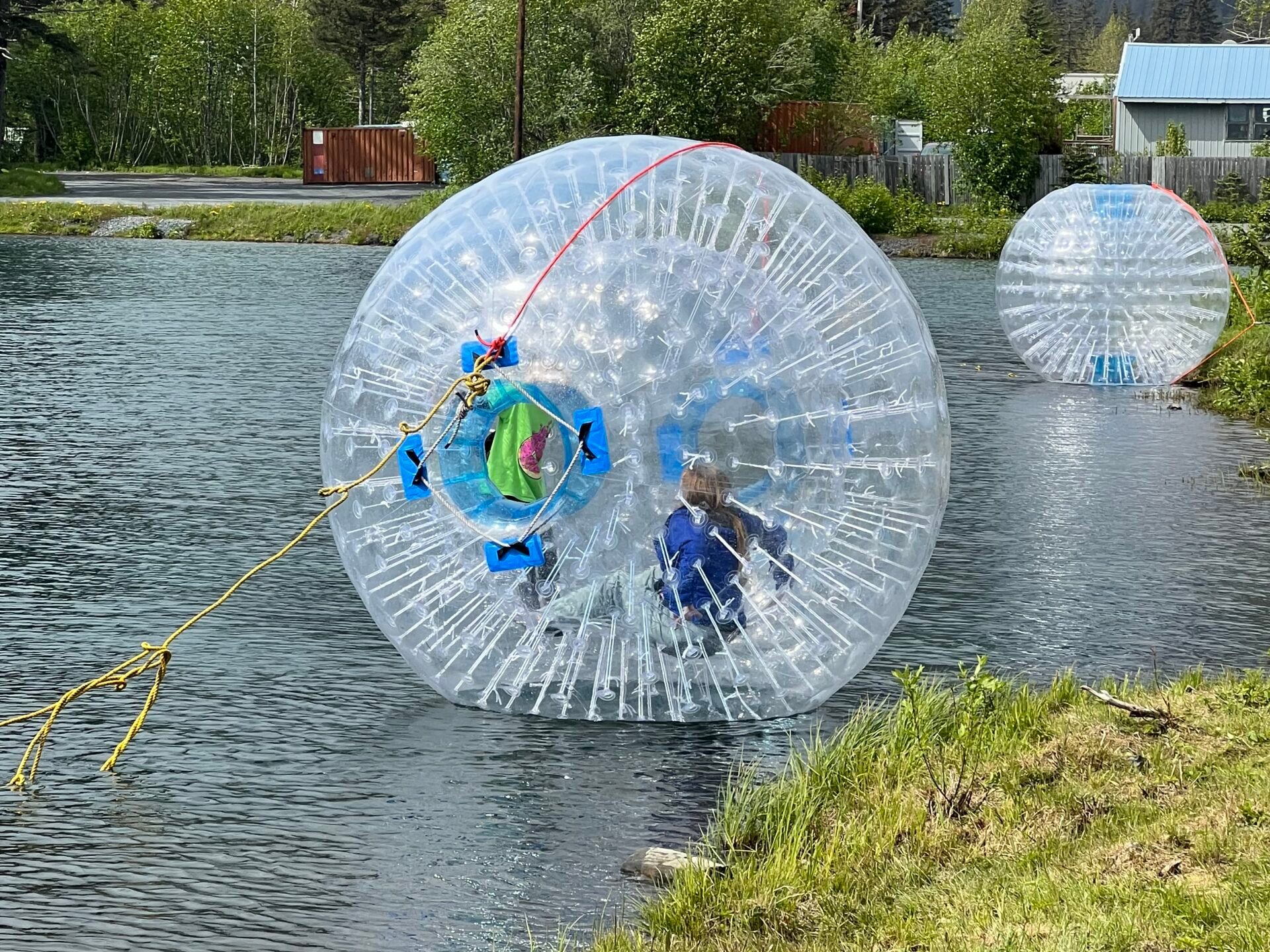 Two people inside large inflatable balls on a water surface with mountains in the background.